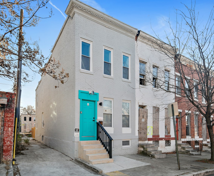 547 N. Pulaski Street after renovation. A light gray Baltimore City corner rowhouse with a teal blue door. Located directly next to a vacant property.