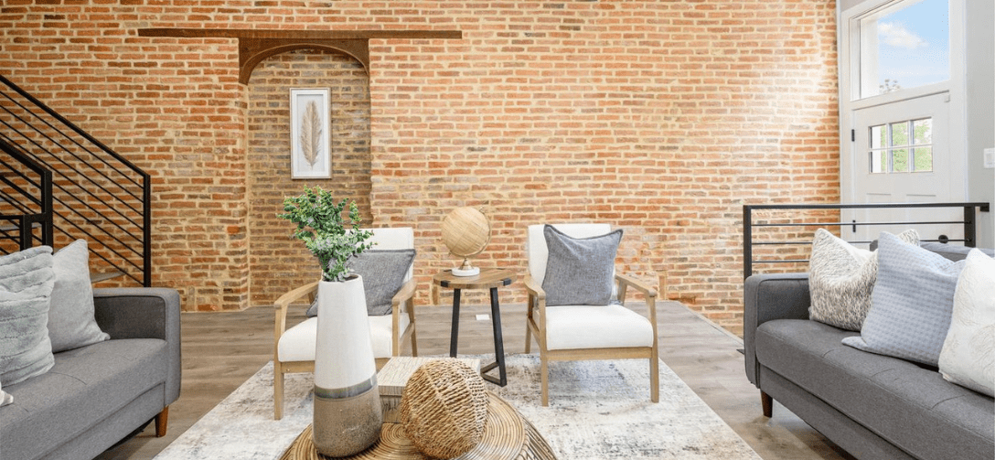 824 Edmondson Avenue interior after renovation. Exposed brick wall with display niche, gray furniture, bright natural lighting coming though window near white door.